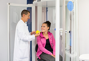 Eine Klinikangestellte spricht mit einer Patientin, die in einer Kabine sitzt, wo ihre Lungenfunktion gemessen wird.