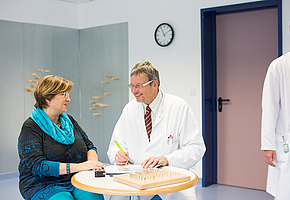 Der Chefarzt der Rehaklinik Heidelberg-Königstuhl sitzt mit einer Patientin an einem Tisch und spricht mit ihr.