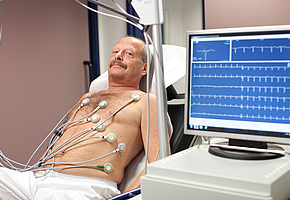 Ein Mann liegt in einem Untersuchungsraum auf einer Liege. Die Aktivität seines Herzens wird anhand eines EKGs aufgezeichnet.